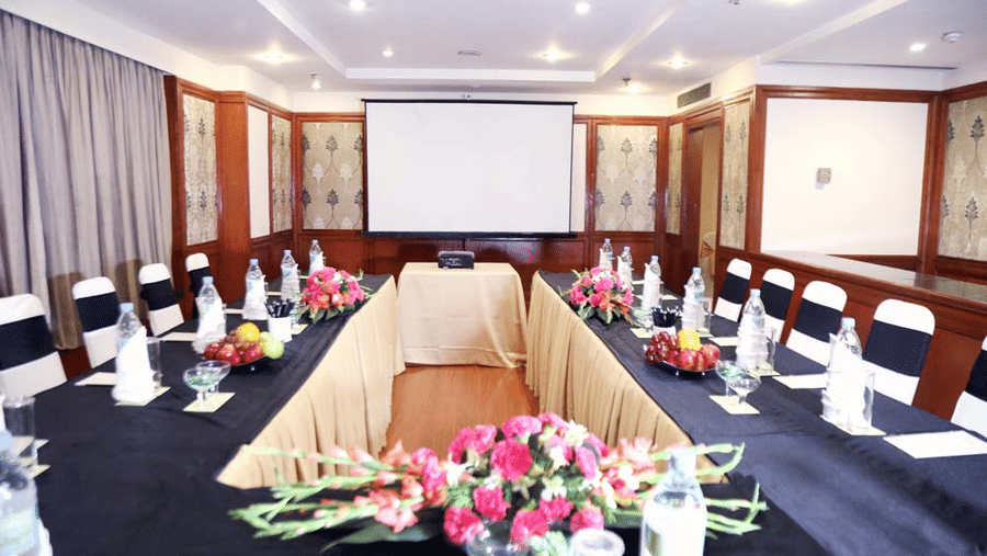 Meeting room at The Bristol Hotel, Gurgaon with tables arranged facing a projector screen, black chair covers, and pink flower arrangements on the tables.