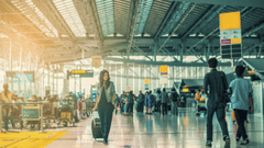 A woman walking through a busy airport terminal while pulling a wheeled suitcase, with travellers, check-in counters, and a spacious glass-roofed concourse in the background.