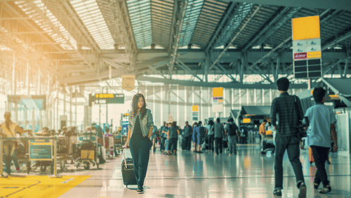 A woman walking through a busy airport terminal while pulling a wheeled suitcase, with travellers, check-in counters, and a spacious glass-roofed concourse in the background.