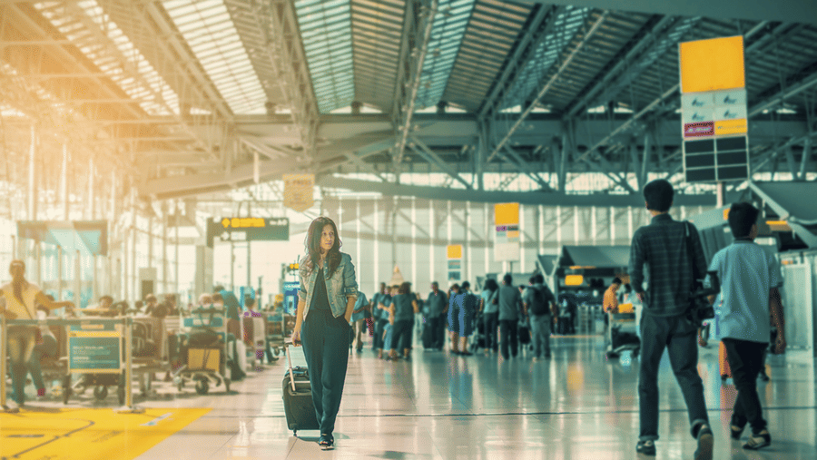 A woman walking through a busy airport terminal while pulling a wheeled suitcase, with travellers, check-in counters, and a spacious glass-roofed concourse in the background.
