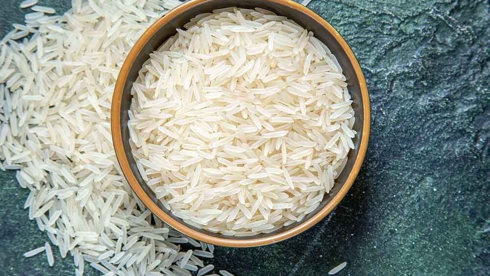 A full bowl of sona masoori rice placed on a dark coloured surface with grains scattered around.