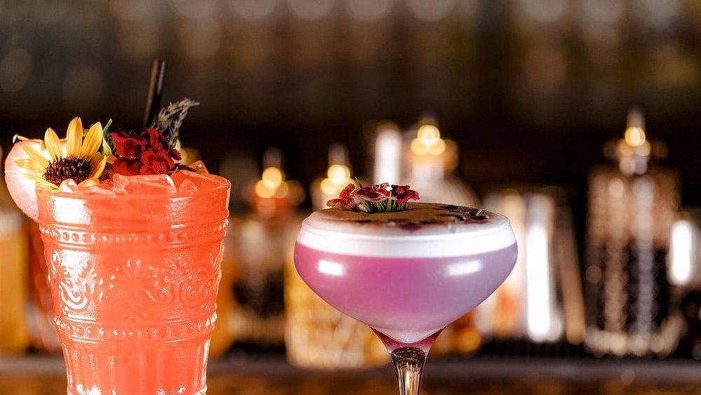 Glass filled with orange beverage topped with a flower accompanied by another glass filled with a lavender purple and pink beverage topped by flowers with a  blurred background.
