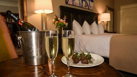 Front view of two champagne glasses filled with champagne , a steel ice bucket and a plate of food on a table in dimly lit room with a neatly made bed and lamps on either side of the bed