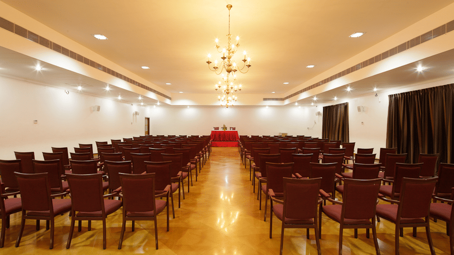 A large conference hall or auditorium with rows of wooden chairs with maroon upholstery facing a small stage under a chandelier | Le Pondy