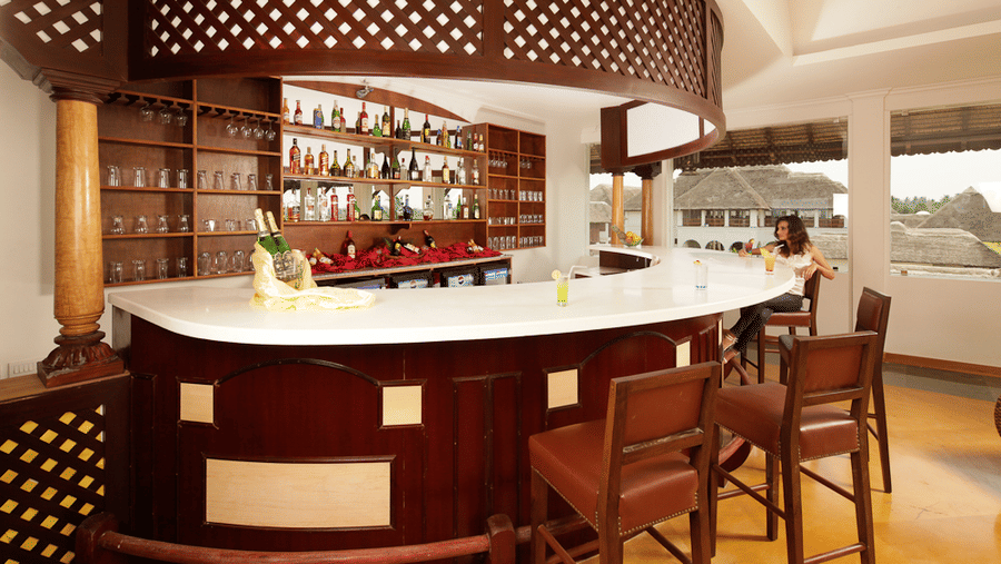 A curved indoor bar counter with a white top and wooden panelling, surrounded by bar stools and backed by shelves of bottles | Le Pondy