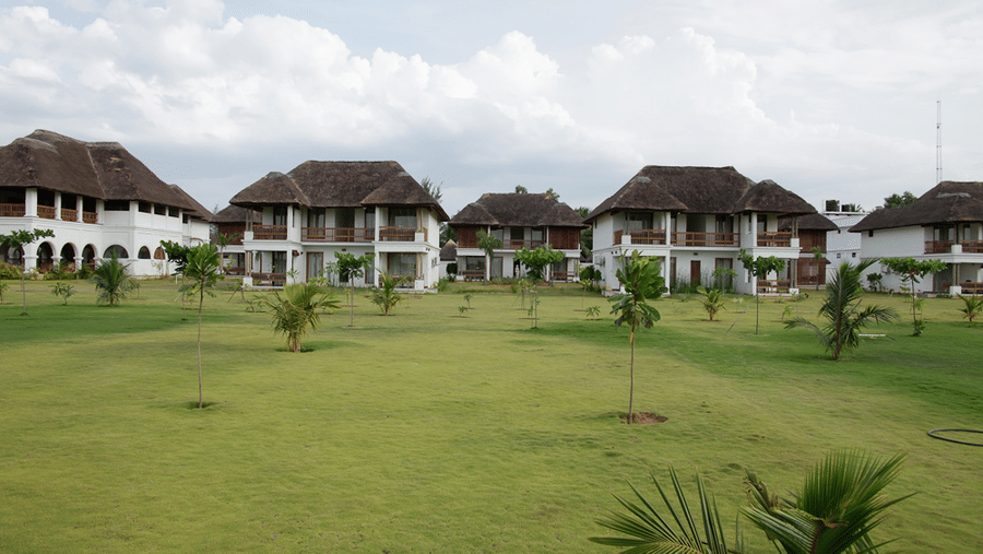 A wide green lawn with young trees situated in front of a row of 2-storey resort villas under a cloudy sky | Le Pondy