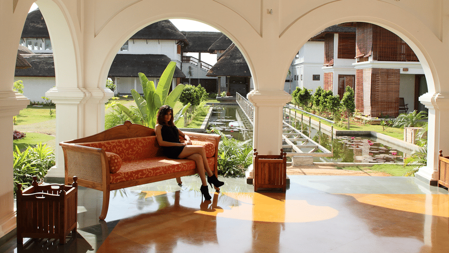 A woman sitting on a wooden sofa with orange cushions in an open-sided resort lounge featuring white arches overlooking a garden | Le Pondy