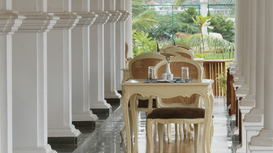 A set of white wooden dining chairs and a table arranged on a veranda behind a row of white columns | Le Pondy