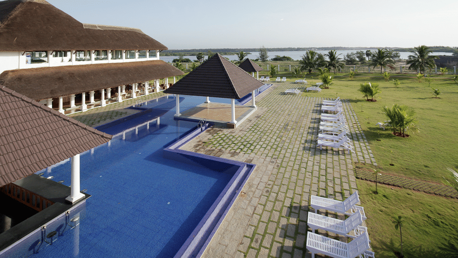 A high-angle view of a blue swimming pool area with a central pavilion, paved patio, rows of sun loungers, and lawns | Le Pondy