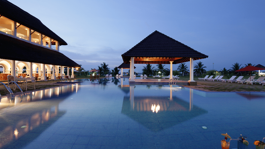 An outdoor swimming pool reflecting a lit pavilion and resort building under a twilight sky with cocktails in the foreground | Le Pondy