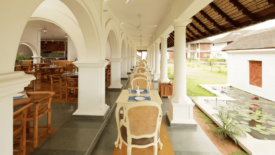 A long outdoor covered restaurant veranda with white arched columns, featuring dining tables and chairs overlooking a garden area | Le Pondy