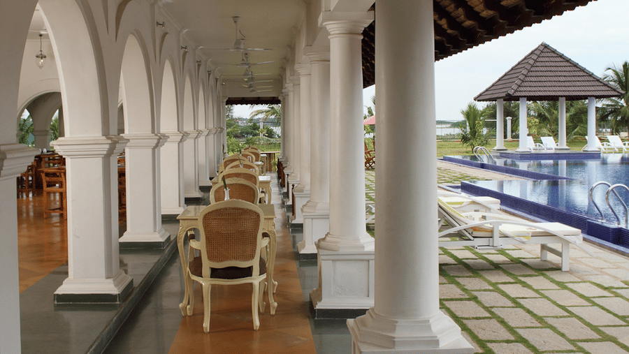 A covered outdoor restaurant veranda lined with white pillars, featuring dining tables and chairs next to a paved pool deck | Le Pondy