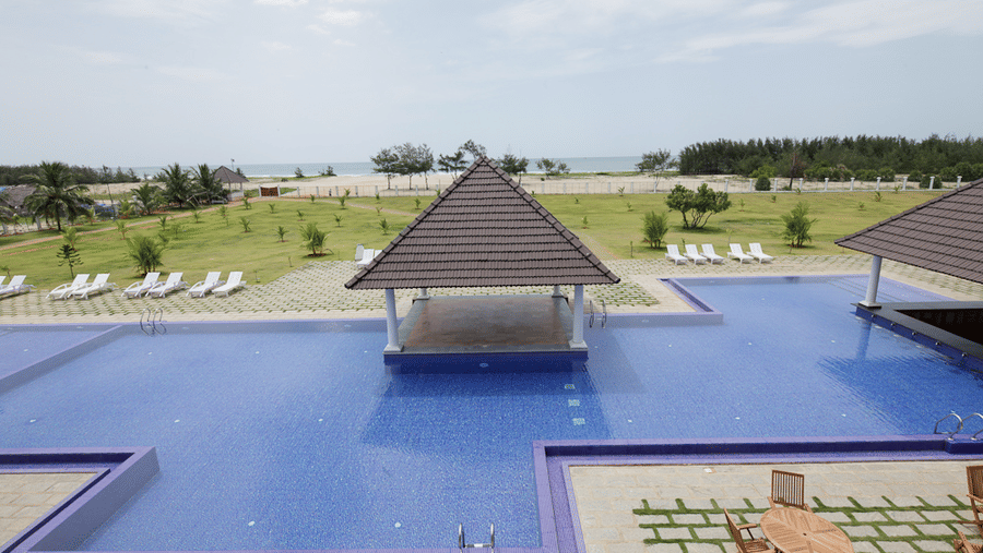 An outdoor blue-tiled swimming pool with a central pavilion, sun loungers, and a view towards a sandy beach area | Le Pondy