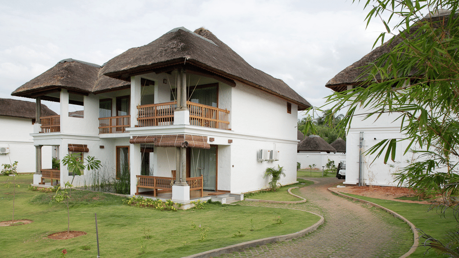 An exterior view of a white 2-storey resort villa with wooden balconies and a thatched roof next to a pathway | Le Pondy