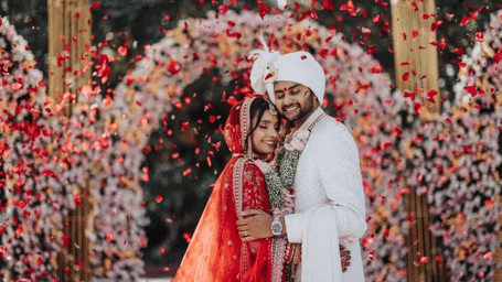 A bride and groom in traditional wedding attire stand under a floral arch.