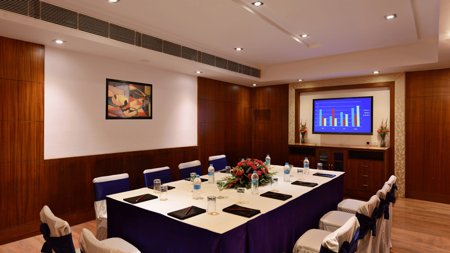 Board room with royal blue seating arrangement, bright lighting, flat screen TV, and wooden flooring.