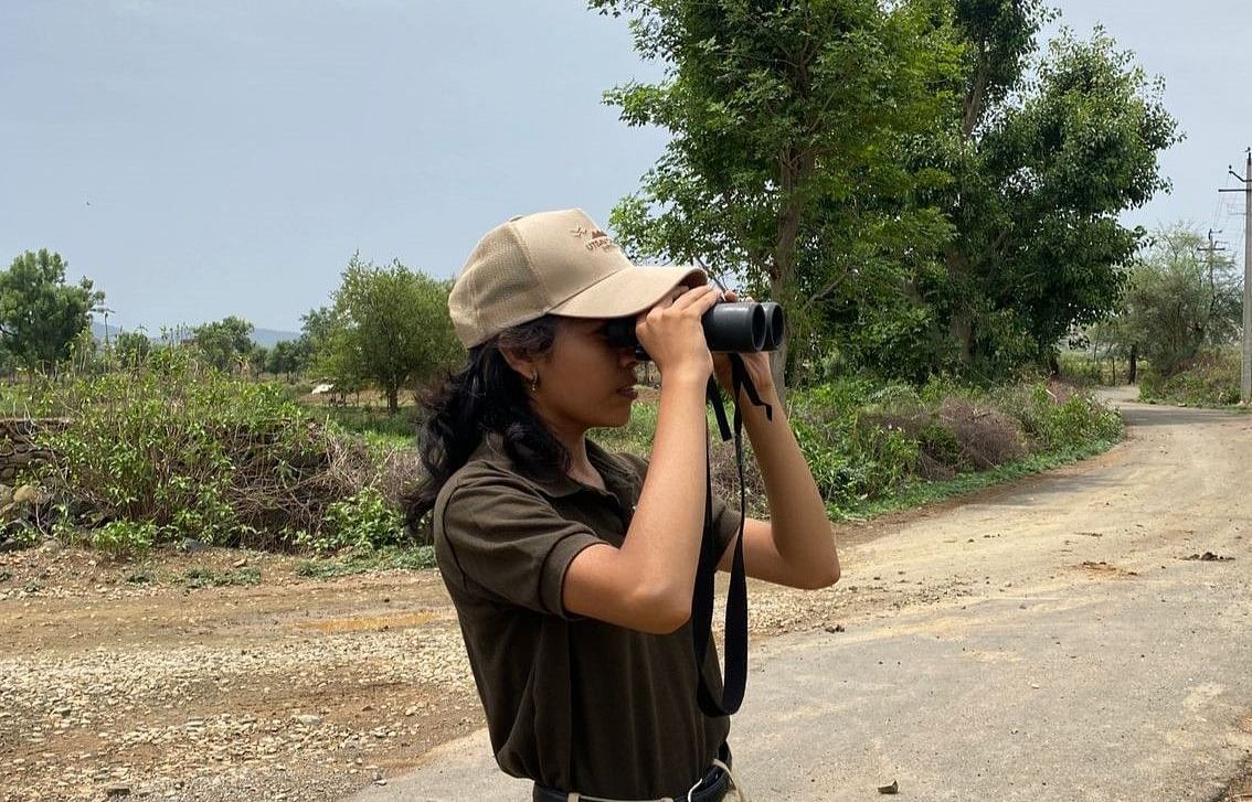 Woman in safari uniform using binoculars on a rural path with trees in the background