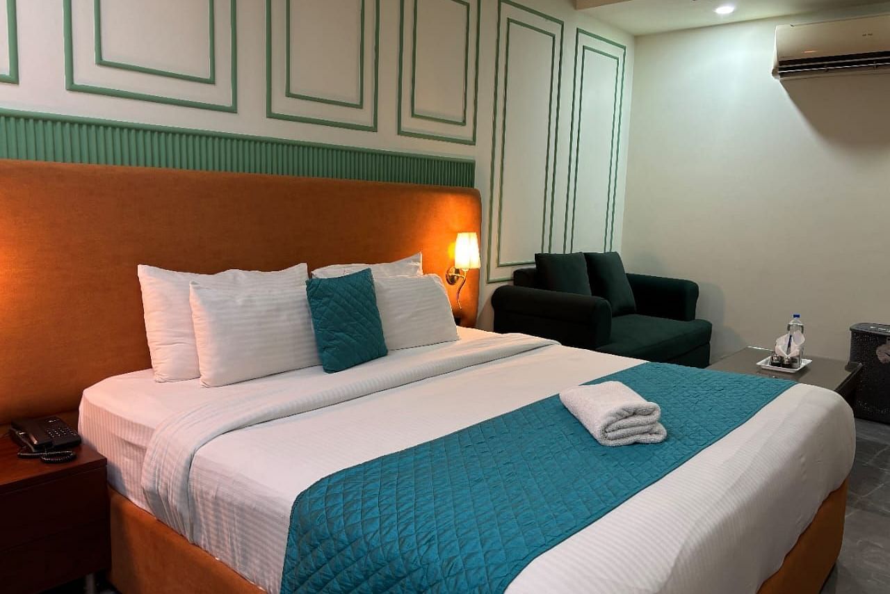 A well-lit room with a double bed dressed in white and blue, and a dark armchair - Asian Suites IFFCO Metro, Gurgaon