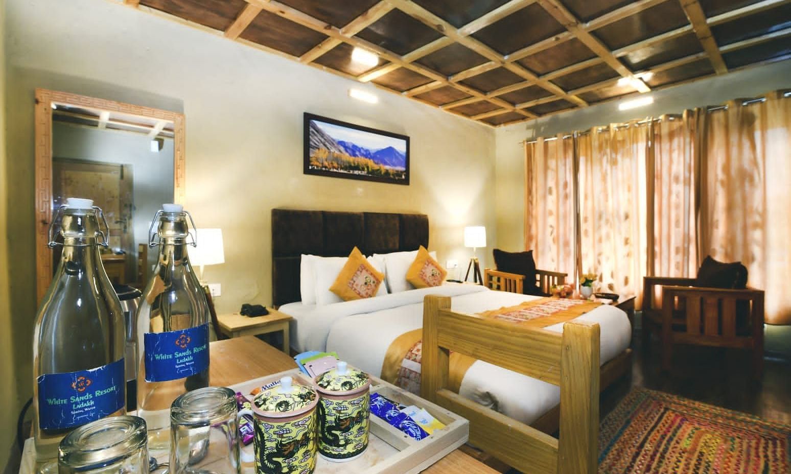 Standard Cottage with a king size bed, a seating area, a wooden ceiling, and bottles and glasses on a table at White Sands Resort, Ladakh.