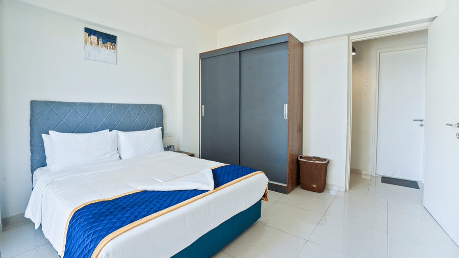 A simple, clean hotel bedroom with a double bed, blue decorative runner, a dark grey wardrobe, and a white door in a minimalist room at Apartel by Aarin, Oragadam.
