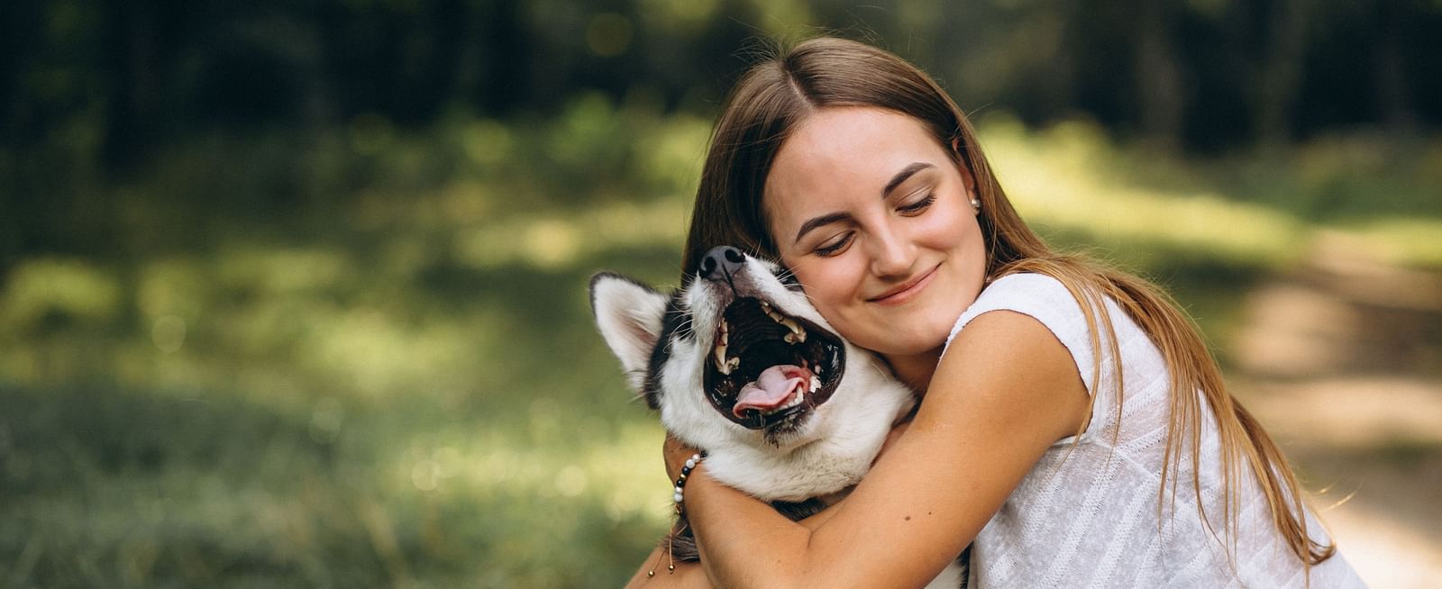A woman with long brown hair and closed eyes hugs a Siberian Husky puppy with its mouth open in a grassy outdoor setting.