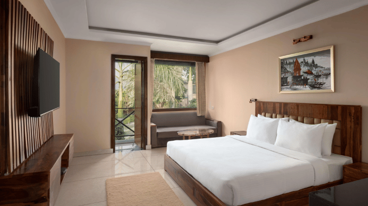 A Bay Room at Corbett Adventure Resort, Ramnagar, featuring a spacious bedroom with a king size bed, comfortable seating area, television, and large windows opening to lush outdoor views.
