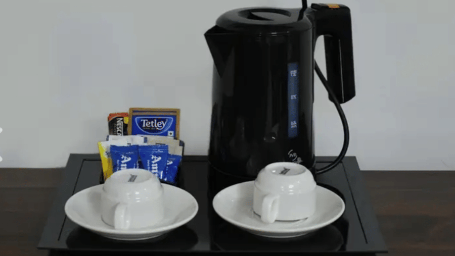 An in-room coffee and tea station at Estherea Bagh, Ranthambore, featuring a black electric kettle, 2 ceramic mugs, and assorted sachets.