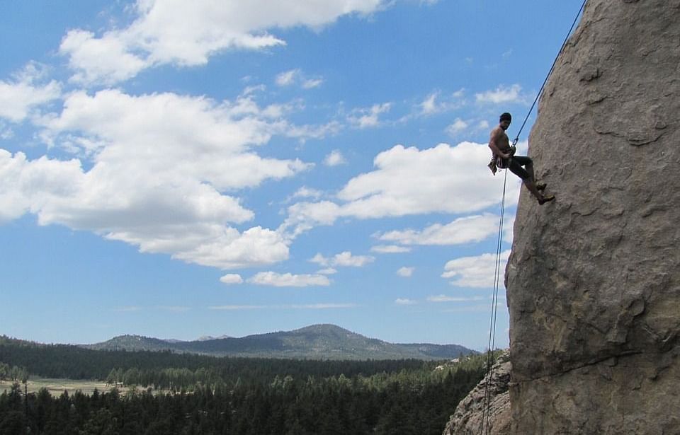 picture of a person rock climbing a tall rock face with the mountains and forest in the background.