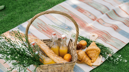 A picnic blanket with a hat and a basket of food laid out on a vibrant green lawn.