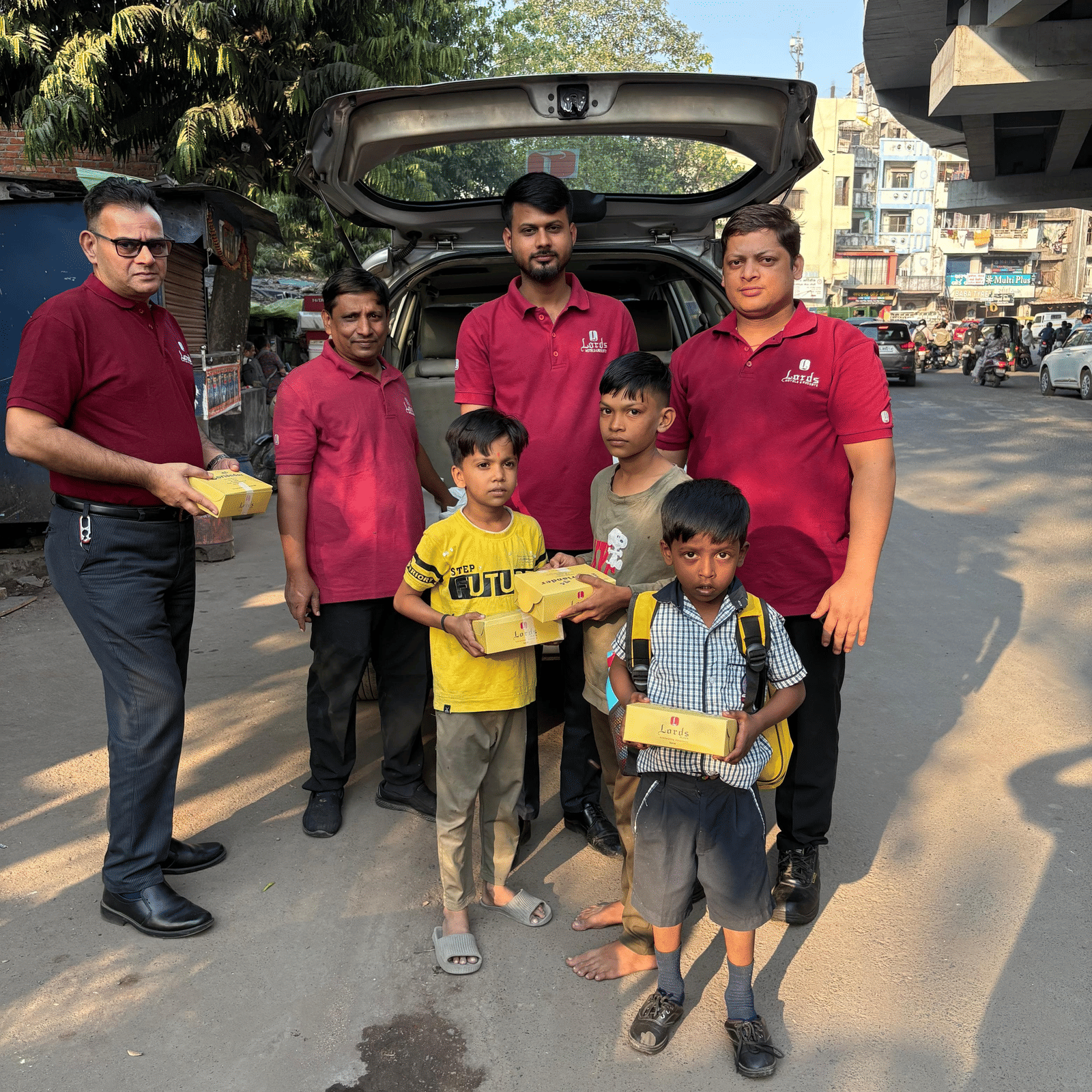 Men in red uniforms distributing packed food to children beside a parked car on a busy street during a community support activity.