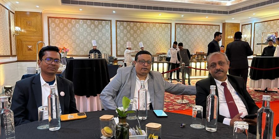 Three men in formal attire seated at a table in a banquet hall with chefs and guests in the background.