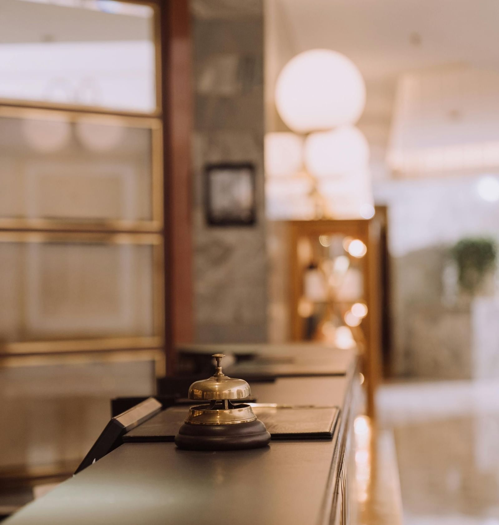 A minimalist interior of a reception with a light-coloured counter, a bell, and a blurred background showing a doorway and a light fixture.