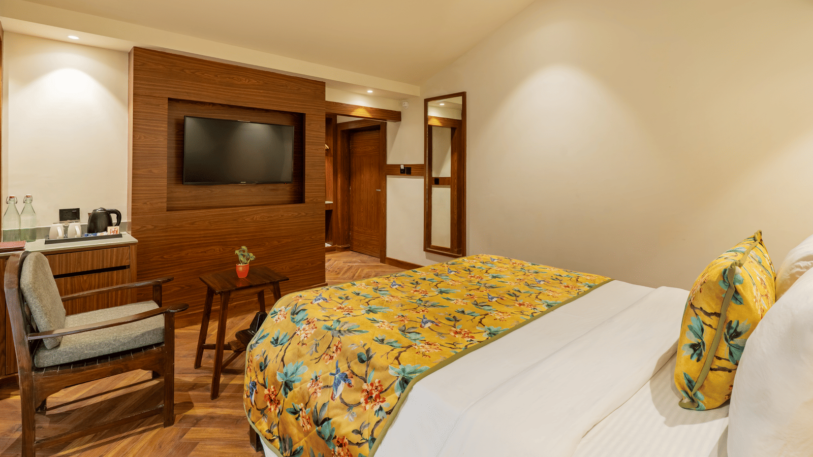 A well-lit spacious bedroom with beige walls and an attached bathroom, featuring a bed facing a television unit, a wooden chair and a table, a cabinet with a small tea station, and a mirror on a wall | Summit by the Ganges Beach Resort & Spa, Rishikesh