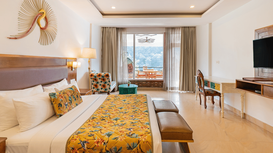A bedroom with a bed, a television, a writing desk with a chair, and an attached balcony overlooking the trees outside | Summit by the Ganges Beach Resort & Spa, Rishikesh