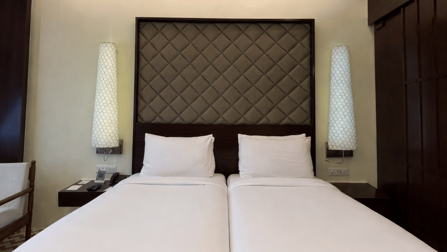 Twin beds with tall, patterned headboards and white linens beneath a ceiling fan in the Superior Twin room at Clarks Amer, Jaipur.