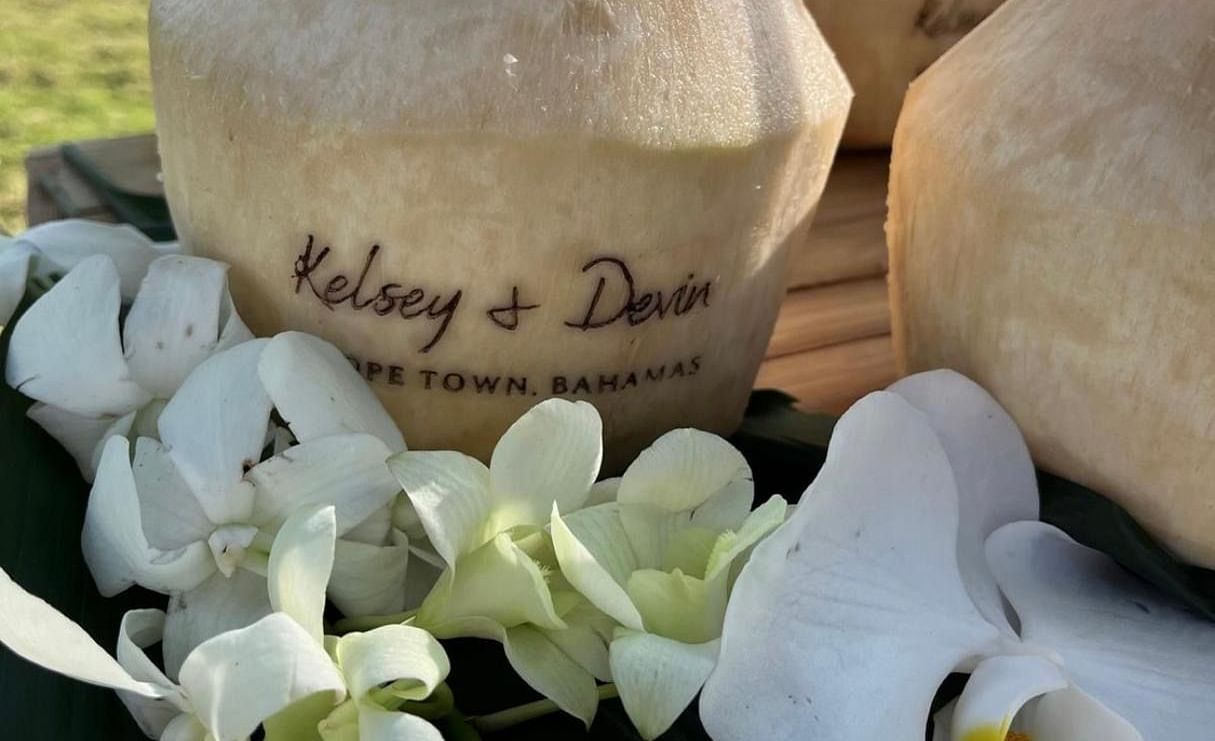Coconuts with straws dipped in are placed on a bamboo table near the beach side. The coconuts have carvings with a couple's name Kelsey and Devin and are placed with orchid flowers. 