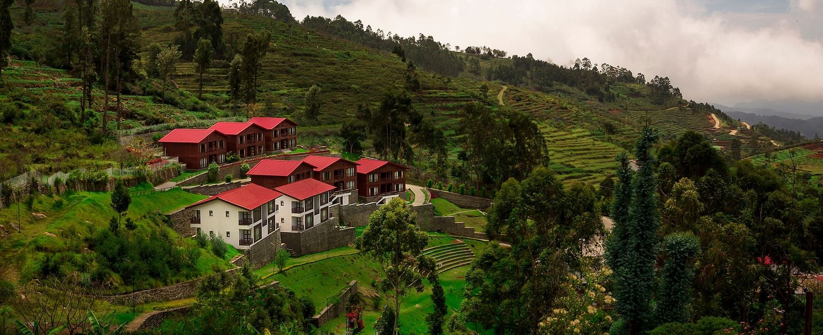 A drone shot of the GReaT Trails Kodaikanal By GRT Hotels property surrounded by lush greenery and trees with clouds hovering over