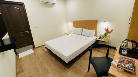A bedroom at Sanctum Suites, Richmond Road, Bangalore features a double bed, a television, a desk with a kettle, a chair, and a dark wood entrance door.