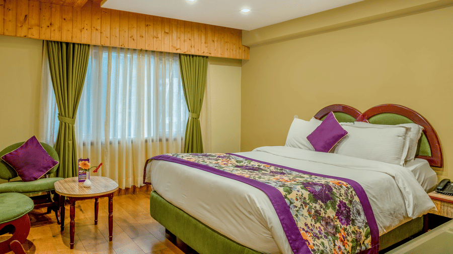 The deluxe plus room with a double bed featuring a padded headboard and decorative bed runner, wooden ceiling panels, bedside tables with lamps, 2 upholstered armchairs with a round coffee table, a tea and coffee setup, and a large window.