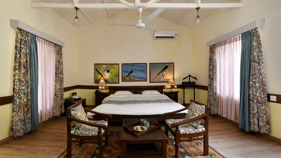 Spacious indoor lounge with high sloped ceiling wooden furniture central table and large windows creating an airy shared space at Neemrana’s Glasshouse on The Ganges suitable for relaxation conversation and quiet leisure indoors