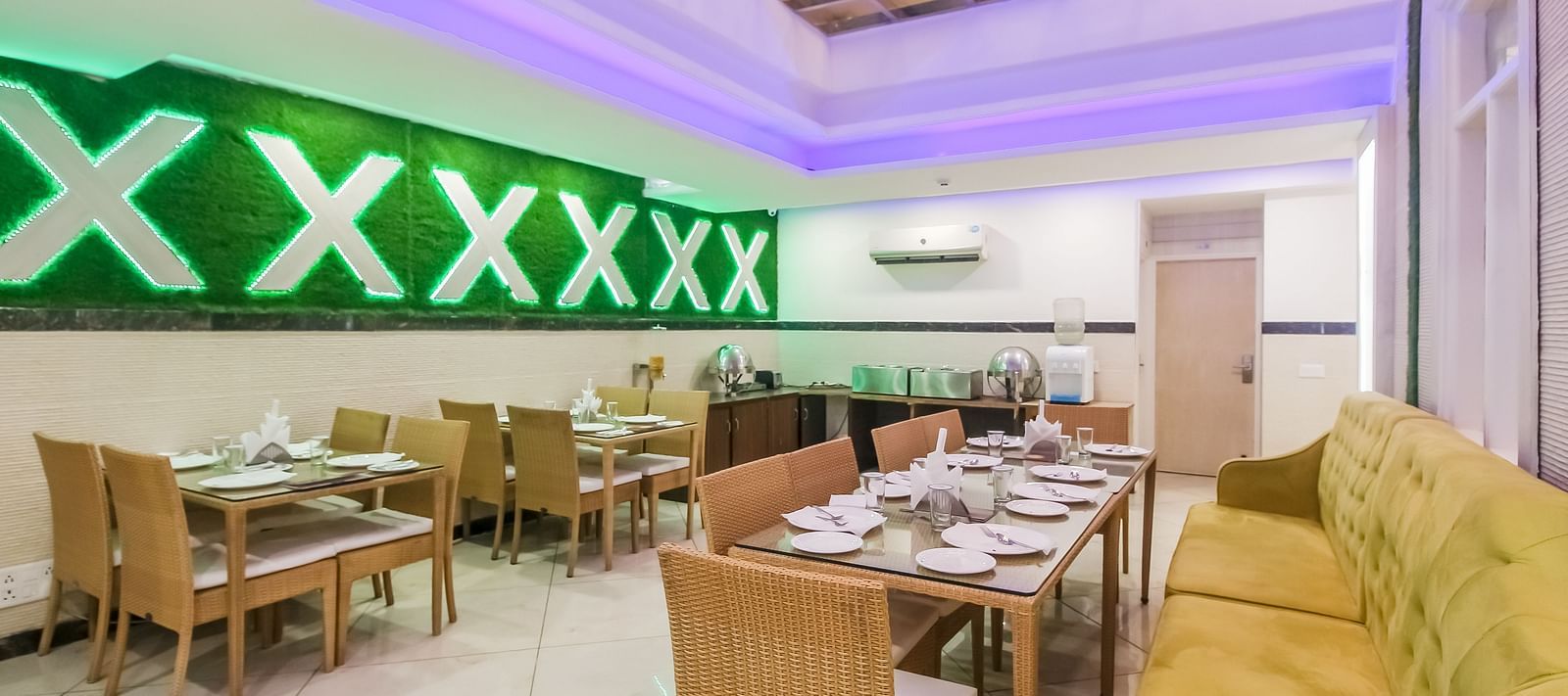 A restaurant interior with a long tufted sofa, wooden tables and chairs, a decorative wall design, and a tiled floor, lit by soft lighting. - Hotel Diamond Plaza, Chandigarh.