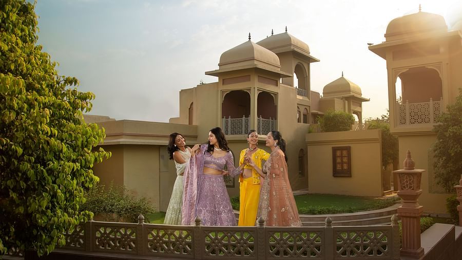 Four women in traditional indian attire dance joyfully in a courtyard of an ornate building, with the early evening sun casting a warm glow.