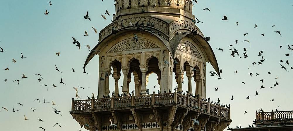 A domed tower with arched openings is part of a larger structure under a clear sky with flying birds.