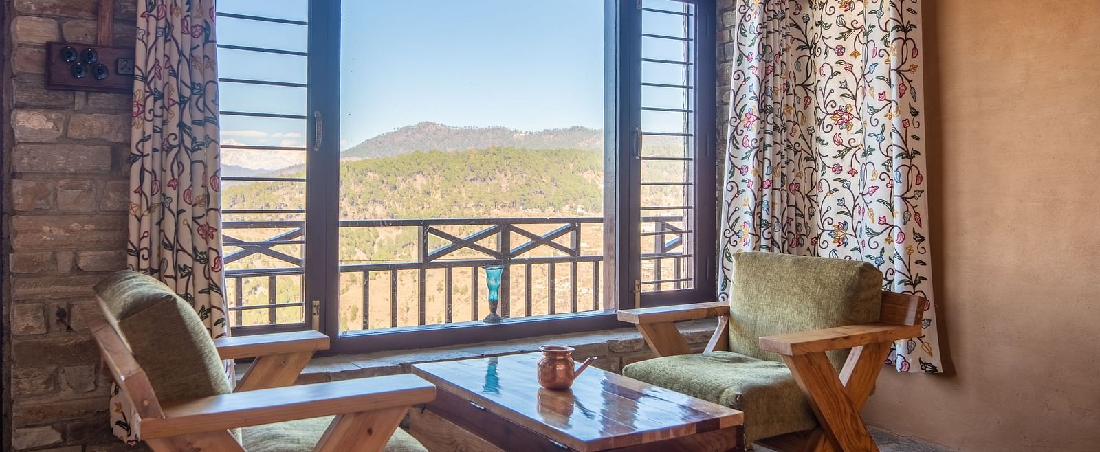 Seating area featuring wooden furniture, floral curtains, and a stunning valley view through a window. - Blusalzz Homes The Himalayan Bungalow, Almora