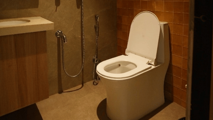 A modern bathroom features a white toilet, a wooden vanity with a sink, a mirror, and a textured wall with a shower fixture at Bougainvillea, Alibaug.