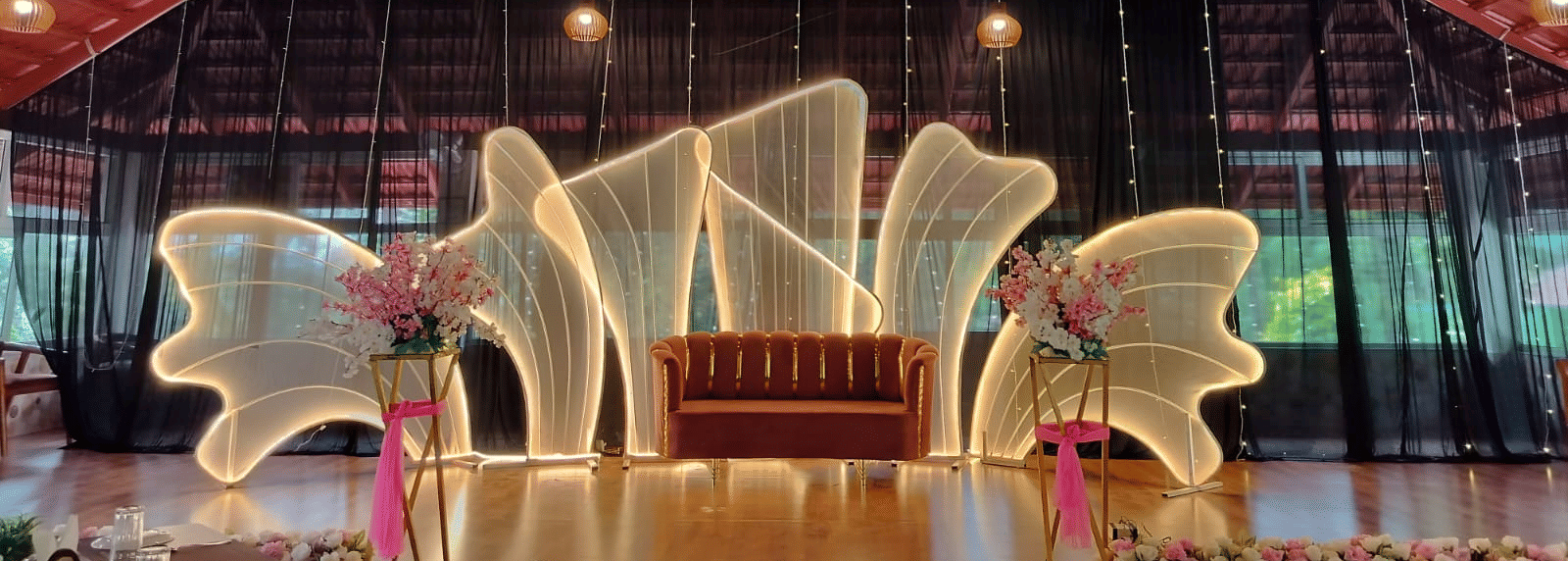 An indoor stage area with a high, exposed truss roof and a large, abstract illuminated white wing-like backdrop at Coorg Orange Blossom Resort and Spa.