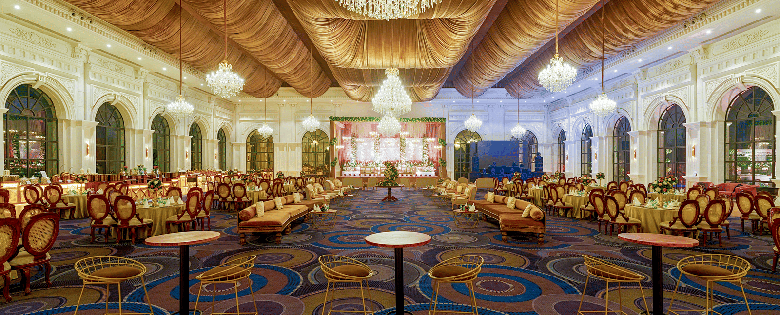 The opulent lobby of Nirvana Luxury Hotel, Ludhiana, showcasing a patterned carpet, comfortable seating areas, elegant chandeliers, and draped golden curtains.