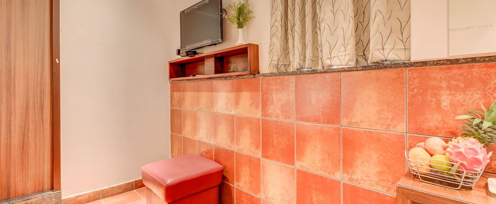 A wall-mounted TV, a cushioned chair, a window covered with curtains, and a table with a fruit basket at Harigovinda's JNStay