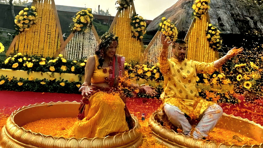 A bride sitting in a shallow pool decorated with marigold flowers during a traditional wedding ceremony at Vedic Village Spa Resort with floral arrangements and greenery surrounding the setting.