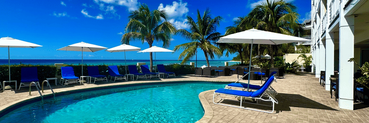 A sunny poolside area surrounded by stone tiles, lounge chairs, umbrellas, and palm trees overlooking the ocean at The Soco Hotel.